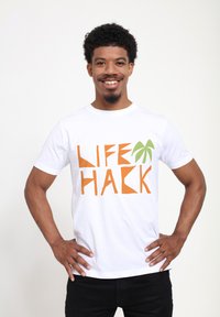 Bílý tričko s výrazným textem "LIFE HACK" v oranžové a zelené barvě, doplněné grafikou palmy. Krátké rukávy a klasický krční výstřih.