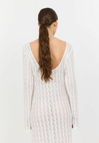 Vit långärmad klänning i spets med V-design i ryggen och vågmönster. Tyg med genomskinlig textur, figurnära silhuett och högslitsar på sidorna.