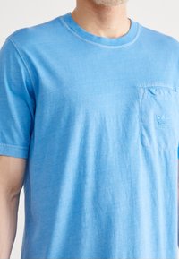 Světle modré bavlněné tričko s kulatým výstřihem, krátkými rukávy a náprsní kapsou s vyšitým logem. Měkká textura.