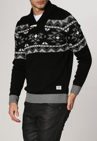 Pull noir en maille avec un motif géométrique gris et un col châle. Présente des poignets et un ourlet côtelés, ainsi qu'une étiquette sur le bas à l'avant.