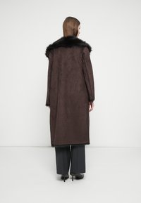 Καφέ σουέτ παλτό με μαύρο τεχνητό γούνινο γιακά, μακριά μανίκια και ευθεία σιλουέτα, φορεμένο πάνω από γκρι παντελόνια. Ελάχιστες ορατές λεπτομέρειες.