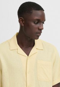 Camicia a maniche corte, con colletto, di lino giallo chiaro, dotata di taschino sul petto, caratterizzata da un tessuto testurizzato e cinque bottoni sul davanti.