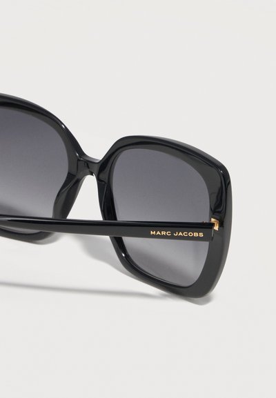 Μαύρα υπερμεγέθη τετράγωνα γυαλιά ηλίου με σκουρούς φακούς και χρυσό κείμενο "Marc Jacobs" στο πλαϊνό, εμφανιζόμενα σε λευκό φόντο.