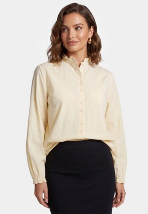 Woman with wavy brown hair wears a cream button-up blouse with ruffled collar and a black fitted skirt, looking slightly to the side.