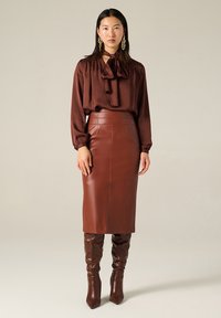 Blusa di satin marrone con colletto annodato, abbinata a una gonna a matita in pelle marrone. Disponibili stivaletti fino al ginocchio con tacco in pelle marrone coordinata.