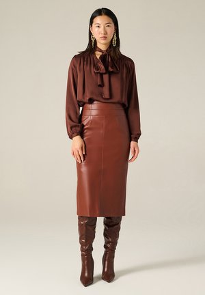 Blusa di satin marrone con colletto annodato, abbinata a una gonna a matita in pelle marrone. Disponibili stivaletti fino al ginocchio con tacco in pelle marrone coordinata.