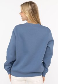 Niebieski bawełniany sweatshirt z okrągłym dekoltem, ściągaczami na rękawach i u dołu. Gładka tekstura i luźny krój, zaprezentowany z tyłu.