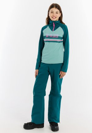 Teal skijack met hoge kraag, rits en grafische print. Teal skibroek met een relaxte fit. Zwarte sneeuwlaarzen met dikke zolen.