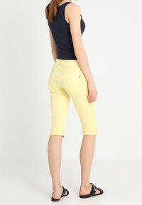 Pantalones cortos amarillos con dobladillo enrollado, hechos de algodón. Combinados con una camiseta sin mangas azul marino y sandalias negras con un detalle de hebilla circular.