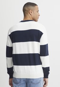 Uomo di profilo che indossa un maglione a righe orizzontali bianco e blu navy e jeans azzurri su uno sfondo bianco uniforme.