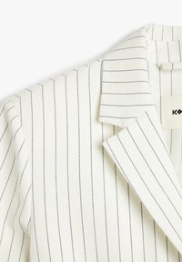 Blazer blanco con finas rayas navy, tejido texturizado, cuello con muesca y solapas bien cortadas. Etiqueta visible en el interior.