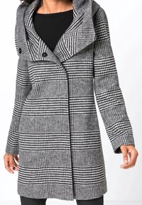 Nainen, jolla on mustavalkoruudullinen keskipitkä takki, jossa on leveä kaulus ja kaksi etunappia, yhdistettynä mustaan toppiin ja housuihin.