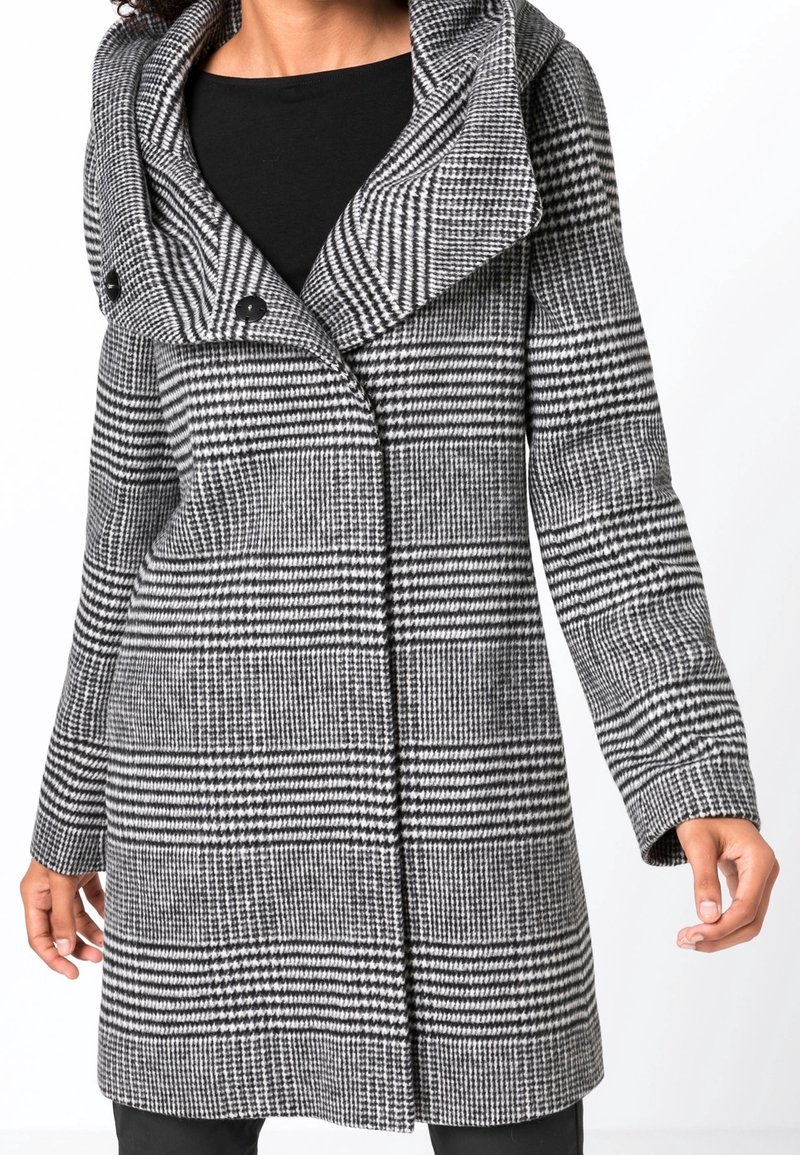 Nainen, jolla on mustavalkoruudullinen keskipitkä takki, jossa on leveä kaulus ja kaksi etunappia, yhdistettynä mustaan toppiin ja housuihin.