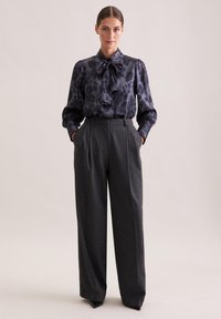 Blusa a pois nera e grigia con dettaglio a fiocco, abbinata a pantaloni grigi antracite a vita alta e gamba larga. Dal taglio sartoriale e design strutturato.