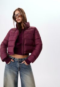 Burgunder pufferjakke med høj krave, lynlås lukning og ribbede manchetter, parret med lyseblå brede jeans. Glat, blankt materiale.