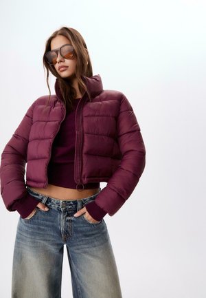 Burgundowa kurtka puchowa z wysokim kołnierzem, zapięciem na zamek i prążkowanymi mankietami, w połączeniu z jasnoniebieskimi szerokimi spodniami dżinsowymi. Gładki, błyszczący materiał.