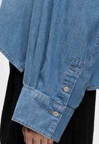 Manche de chemise en jean bleu clair avec deux boutons beiges au poignet, portée sur un vêtement noir, couvrant partiellement une main.