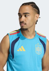 Light blue sleeveless jersey featuring a yellow Adidas logo, navy accents, and Spain's national emblem with a star above. Textured pattern overall.