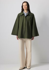 Veste oversize vert olive avec des manches larges, col boutonné et texture lisse, assortie à un pantalon beige à jambes larges et des chaussures claires.