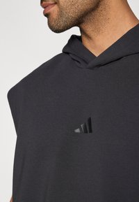 Pilka be rankovių gobtuvinė su minkšta tekstūra. Viršuje gausiai matomas juodas „Adidas“ logotipas ir patogaus dizaino gobtuvas.