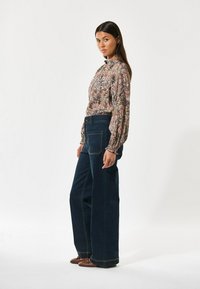 Blusa floreale a maniche lunghe con colletto arricciato; jeans in denim scuro a gamba larga e vita alta; scarpe piatte marroni; modella in piedi di lato.