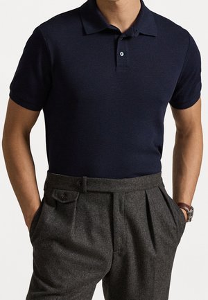 Mann trägt ein marineblaues Poloshirt mit kurzen Ärmeln, das in graue, hoch taillierte, plissierte Hose gesteckt ist, mit den Händen in den Taschen.