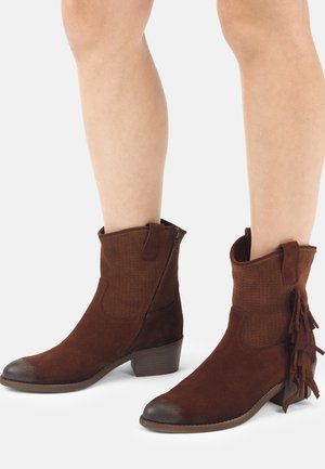 Brune ruskind midt på læggen-støvler med sidelås og frynsedetalje, båret på bare ben med lave stablerede hæle og runde tæer.