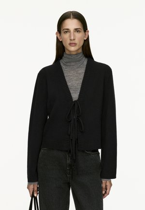Black knitted cardigan with a V-neck, tied front closure, and long sleeves. Detailed with ribbed edges and worn over a gray turtleneck.