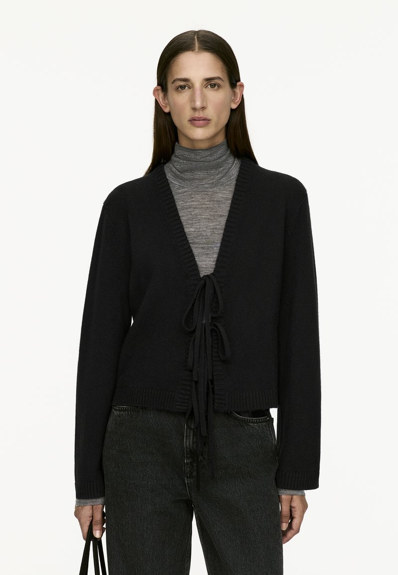 Cardigan noir en tricot avec un col en V, fermeture à nouer à l'avant et manches longues. Détail avec des bords côtelés et porté sur un pull col roulé gris.
