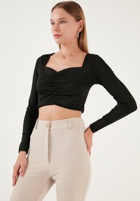 Top nero a maniche lunghe cropped con scollatura quadrata e arricciature frontali, abbinato a pantaloni beige a vita alta con chiusura a bottone.