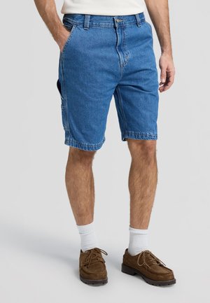 Άνδρας που φοράει μπλε τζιν σορτς με μπροστινές και πλαϊνές τσέπες, λευκές κάλτσες και καφέ δερμάτινα παπούτσια με κορδόνια, στέκεται μπροστά σε ένα απλό φόντο.