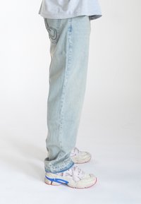 Jeans de mezclilla de un azul claro con un corte relajado, que presentan detalles de bolsillo trasero cosidos. Combinados con zapatillas blancas con acentos en azul y rojo.