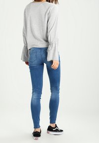 Haut gris à manches longues évasées, associé à un jean skinny bleu et des baskets noires à semelles blanches.