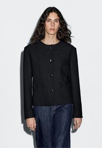 Svart texturerad jacka med rund halsringning, framknappar och två sidofickor. Stylad med mörka denimbyxor.