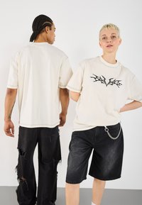 Béžové oversized trička s černým grafickým potiskem. Model vlevo v černých potrhaných kalhotách, vpravo v šedých šortkách s řetězovým akcentem.