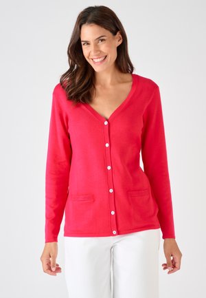 Mujer sonriente, vistiendo un cárdigan rojo brillante abotonado con dos bolsillos delanteros y pantalones blancos, de pie frente a un fondo claro y liso.