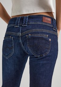 Close-up de uma pessoa a usar jeans de ganga azul escura com bolsos traseiros visíveis, costura e um etiqueta de marca de couro na cintura.