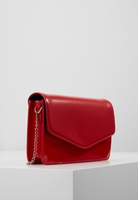 Rød skinnende læderclutch med klaplukning, kæderem og glat overflade. Har et minimalistisk design og skrå kanter.