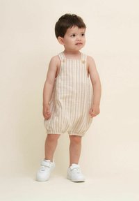 Lettet, stribet beige jumpsuit med justerbare stropper og knapdetaljer, der har samlinger ved benene. Sammen med hvide sneakers.