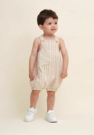 Lettet, stribet beige jumpsuit med justerbare stropper og knapdetaljer, der har samlinger ved benene. Sammen med hvide sneakers.