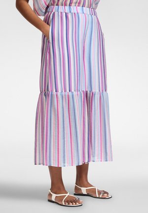 Vrouw draagt een pastelkleurige gestreepte rok tot op de enkels met zakken, gecombineerd met witte platte sandalen, staand op een neutrale achtergrond.