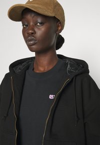 Black zip-up hoodie with a soft texture, highlighted by a pink logo on a black t-shirt underneath. A corduroy tan cap complements the outfit.