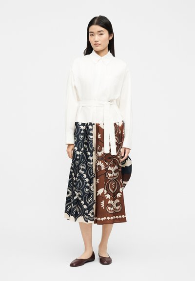 Γυναίκα όρθια με λευκό πουκάμισο με γιακά, λευκή ζώνη και φούστα μήκους μέσης με ναυτικό και καφέ ανθικό μοτίβο, σε συνδυασμό με σκοτεινά φλατ παπούτσια.