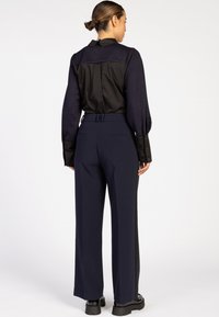 Maßgeschneiderte schwarze Bluse mit Kragen, langen Ärmeln und Knopfmanschetten, kombiniert mit navyblauen, hoch taillierten weiten Hosen.