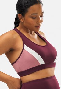 Sujetador deportivo burdeos con paneles en contraste en rosa y blanco, hecho de una tela suave. Presenta un escote en forma de bote y un diseño de espalda nadadora.