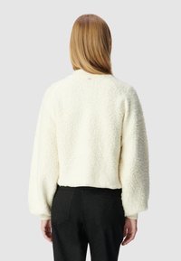 Pull cropped fluffy crème avec des manches ballon et des poignets côtelés. Porté sur un pantalon noir texturé, montrant une coupe décontractée et une texture douce.