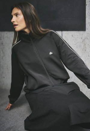 Čierna bunda na zips s kontrastnými bielymi pruhmi, vyrobená z hladkej, elastickej tkaniny; nosená s dlhou čiernou sukňou, s minimálnym logom.