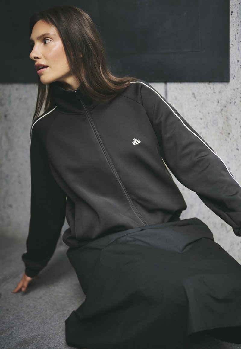 Črna zip-up jakna z kontrastnimi belimi črtami, izdelana iz gladkega, elastičnega materiala; nosi se z dolgo črno krilo, minimalni logotip.