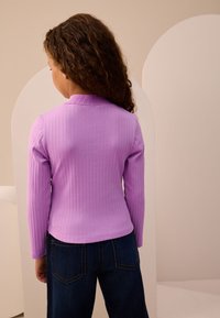 Lilla ribbet langærmet top med høj krave, kombineret med mørkeblå jeans. Stoffet virker blødt og tætsiddende med en subtil tekstur.