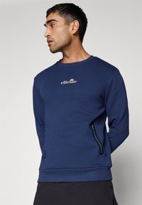 Granatowy sweter z okrągłym dekoltem, długimi rękawami, bocznymi kieszeniami na zamek i małym logo na piersi. Gładka tekstura tkaniny.